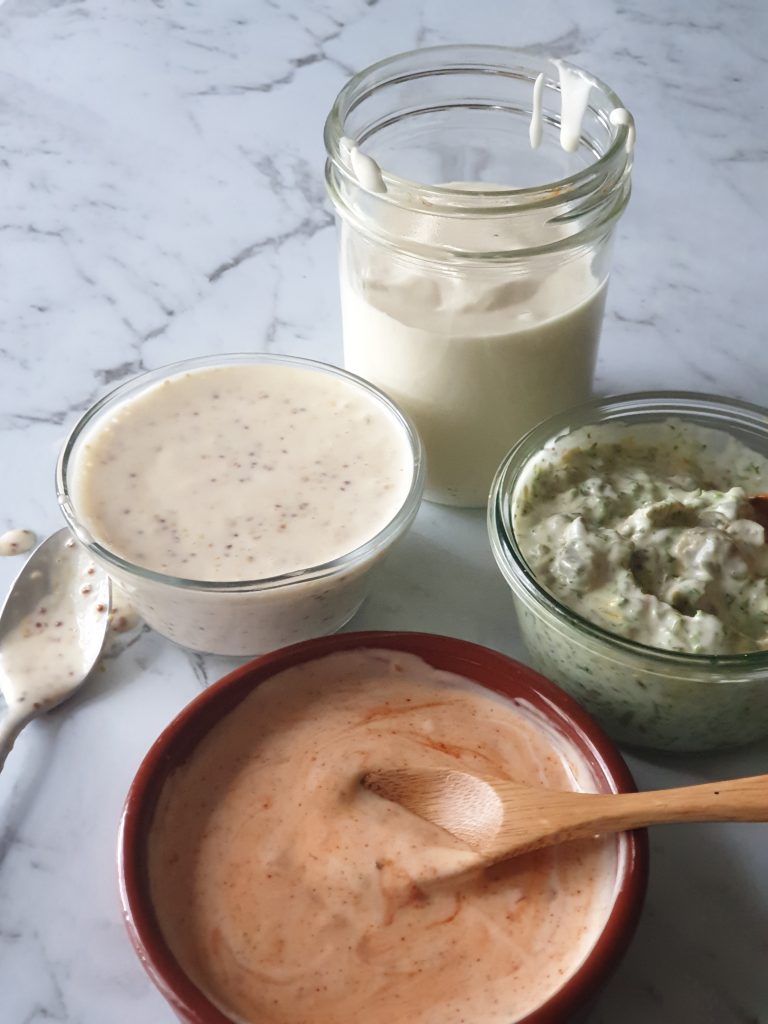 Jar of aquafaba mayo with 3 bowls of flavoured mayo on marble bench