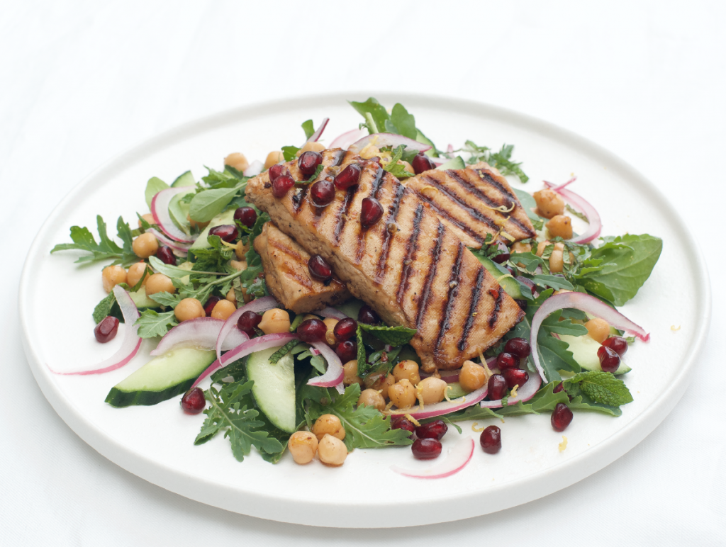 white plate with char grilled slices of tofu on a rocket, chickpea, cucumber and pickled red onion salad with garnish of mint and pomegranate seeds