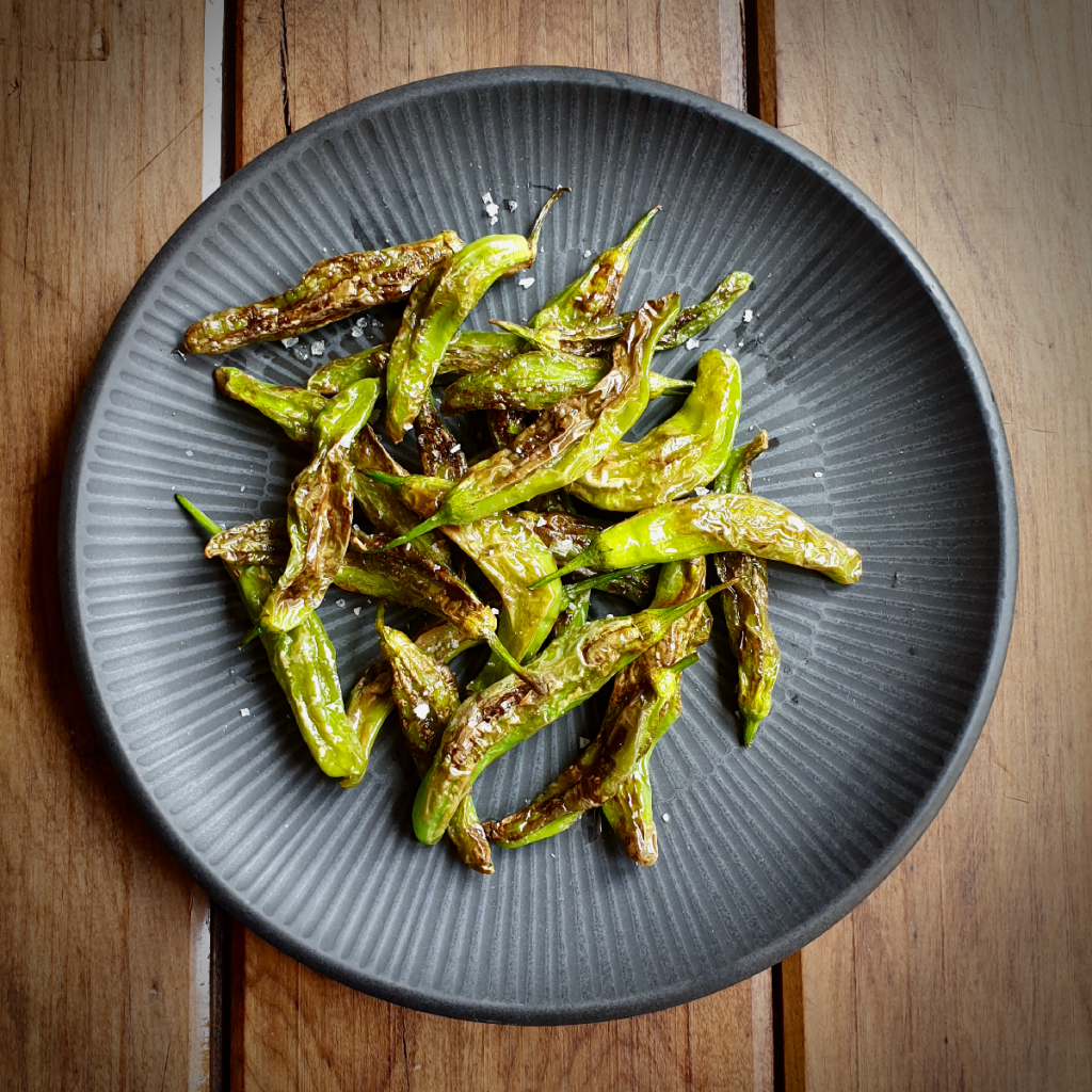 a dark plate with blistered shishito peppers on a wood slat table