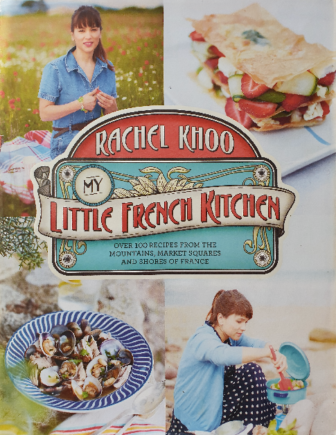 The cookbook cover features a montage of images around the title "My Little French Kitchen" by Rachel Khoo. At the top, Rachel Khoo is seen in a denim shirt with a natural backdrop, smiling at the camera. The central image is a decorative banner with the title, styled in ornate lettering typical of French aesthetics, with the subtitle "Over 100 recipes from the mountains, market squares and shores of France" below it. Below the banner, there's a photograph of a dish with escargots, and in the bottom right corner, Rachel Khoo is cooking, engaged in her craft. The images suggest a personal journey through French cuisine, highlighting its diversity and the author's hands-on approach.