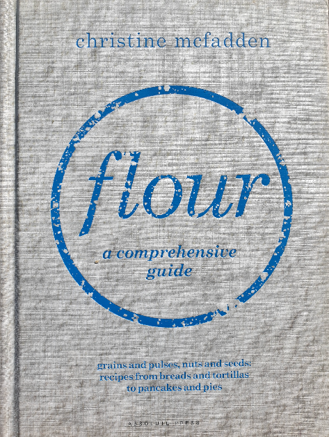 Cover of the cookbook of the month 'Flour: A Comprehensive Guide' by Christine McFadden, featuring a textured light grey background with a large blue circular stamp-like design that reads 'flour' in the center. The author's name and the subtitle 'grains and pulses, nuts and seeds: recipes from breads and tortillas to pancakes and pies' are also displayed, emphasizing the book's focus on diverse flour options.