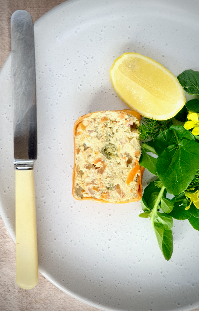 This image features a beautifully plated vegan smoked salmon pâté, served as a slice on a textured white plate, garnished with a fresh wedge of lemon and a watercress, herbs and edible flowers. The pâté has a creamy, flaky texture, with visible herbs and what may be pieces of carrot salmon, red onion and capers. A classic butter knife with a cream handle lies adjacent to the plate, complementing the elegant and fresh presentation. This setup suggests a sophisticated serving suggestion for a brunch or light lunch, emphasising the pâté's versatility and appeal in a fine dining context.