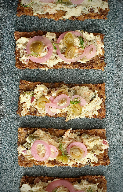 This image showcases an appetizing display of vegan smoked salmon pâté served on slices of crisp multigrain bread. Each piece is elegantly topped with pickled red onions and capers, adding vibrant color and texture contrasts. The pâté is chunky, with visible flakes that mimic the texture of traditional salmon pâté, garnished with a sprinkle of fresh herbs that likely enhance the flavor. The arrangement is meticulous and symmetrical, emphasizing the care in presentation, suitable for a sophisticated appetizer or a part of a gourmet vegan spread.