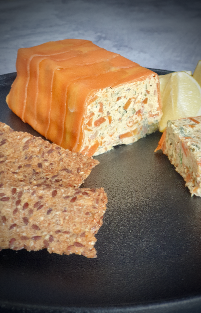 This image showcases a beautifully presented vegan smoked salmon pâté served in a terrine form. The pâté has a salmon-like appearance, wrapped in what looks like smoked salmon, which is likely achieved using thinly sliced carrots It's paired with lemon wedges and crispbread, which adds a nice contrast of textures. The pâté itself seems to be packed with finely chopped herbs , red onion and capers adding freshness and a hint of spice to the dish.