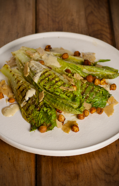 A close-up of a white plate showcasing Vegan Grilled Lettuce Caesar Salad. The salad includes grilled romaine lettuce hearts, roasted chickpeas with seaweed, drizzled with a creamy dressing, and garnished with plant-based parmesan shards. The plate is set on a wooden surface.