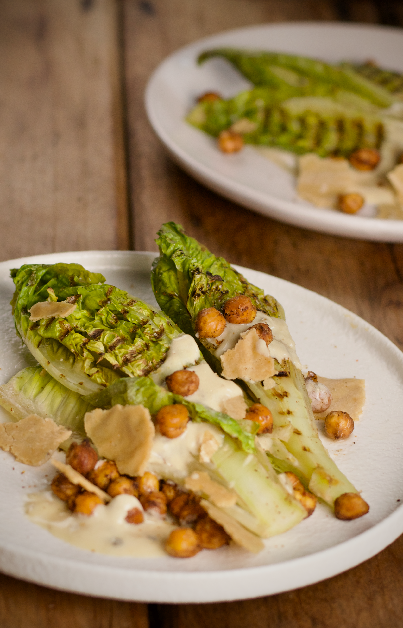 2 white plates with Vegan Grilled Lettuce Caesar Salad, featuring grilled lettuce, roasted chickpeas seasoned with seaweed, creamy dressing, and shards of plant-based parmesan cheese. The dish is presented on a rustic wooden table.