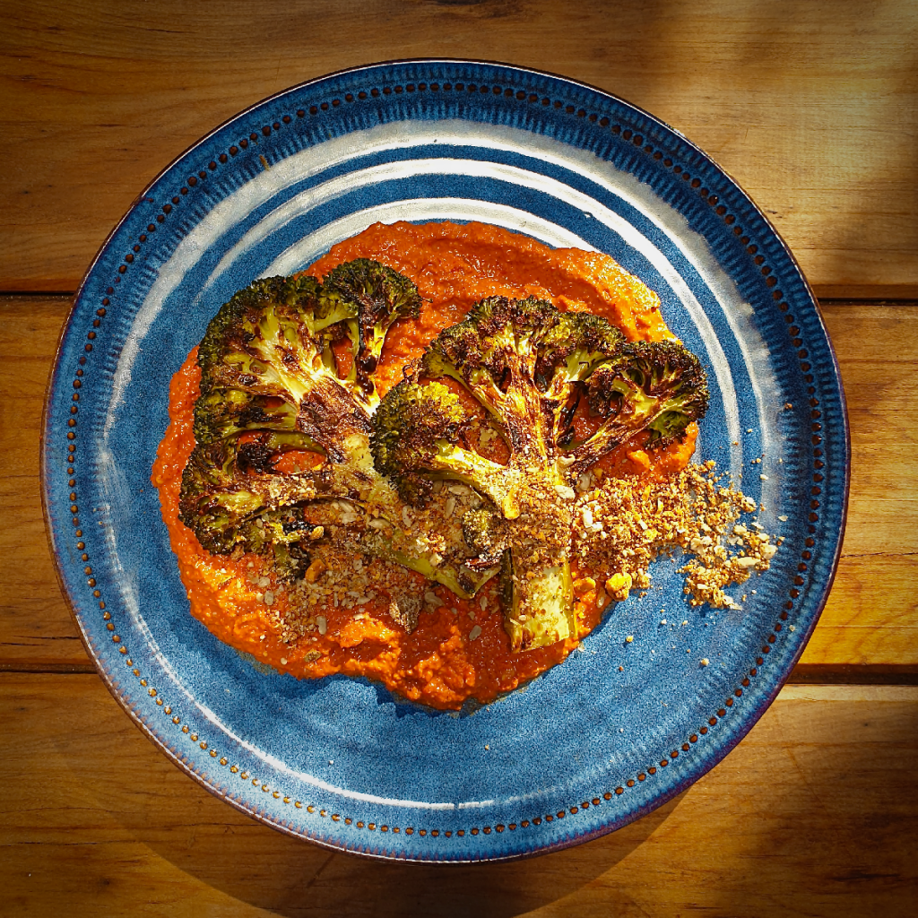 roasted and charred broccoli steaks on a bed of brick red muhammara with a garnish of dukkah on a blue plate set on a rustic wooden board.