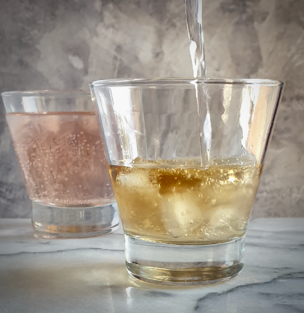 two glasses of homemade violet colour changing gin. One pink, the other with tonic being poured into the amber coloured gin which will turn pink.