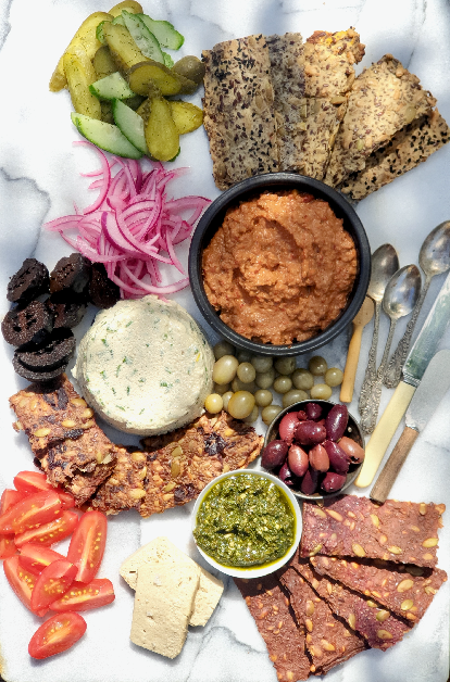 a marble platter with a selection of easy whole-food gluten -free crackers, homemade plant based cheeses, fresh tomatoes, olives, green tomatoes capers, pickled walnuts slices and red pickled onions and a plant-based pesto with vintage spoons and knives for serving