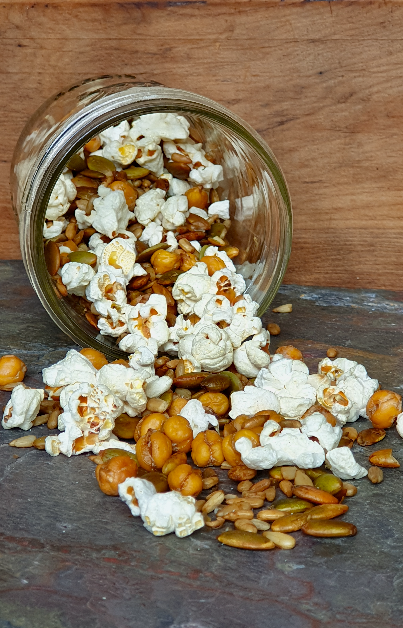 A glass jar tipped over on a slate surface, spilling out an allergy-friendly trail mix of popcorn, roasted chickpeas, pumpkin seeds, and sunflower seeds, set against a wooden backdrop