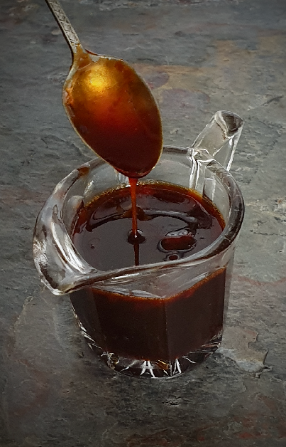 A spoon drizzling thick, dark watermelon molasses into a glass jug on a slate background