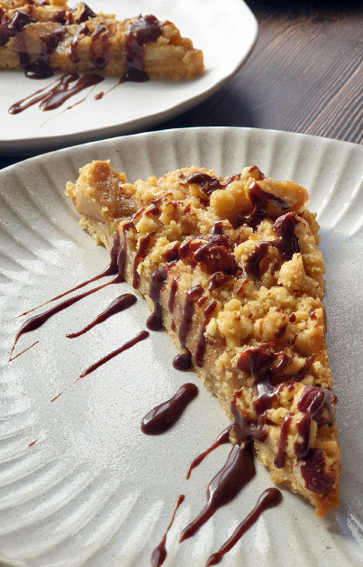 Close-up of a single slice of pear and caramel oat slice with hazelnut crumble, drizzled with chocolate caramel sauce, served on a cream-coloured plate with more slices in the background