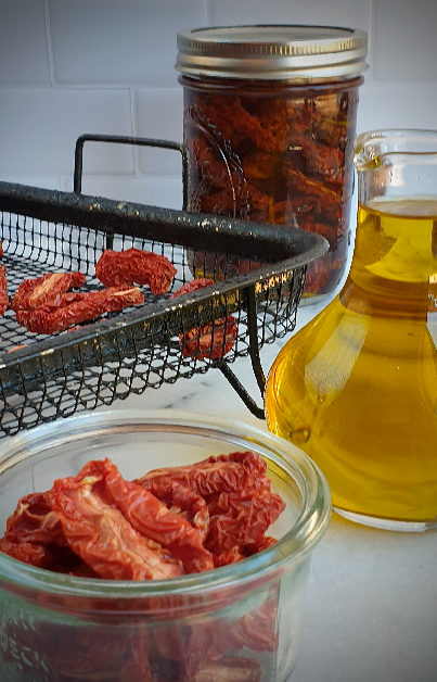Close-up of semi-dried tomatoes in a small glass dish, with a wire rack of drying tomatoes behind them, a jar of tomatoes in oil, and a small glass bottle of golden olive oil on a kitchen bench.